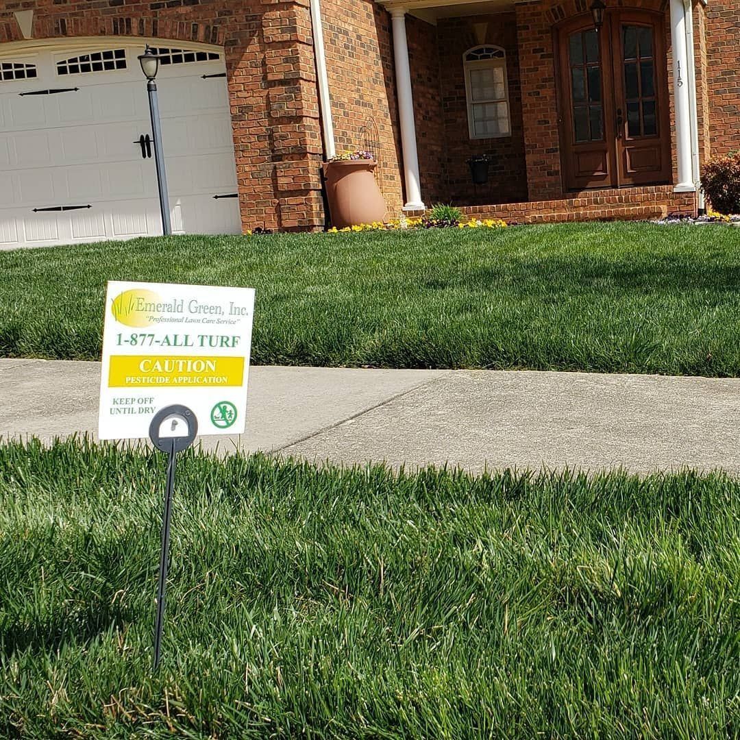 A green lawn with a lawn care sign in front of a brick house with a white garage door.