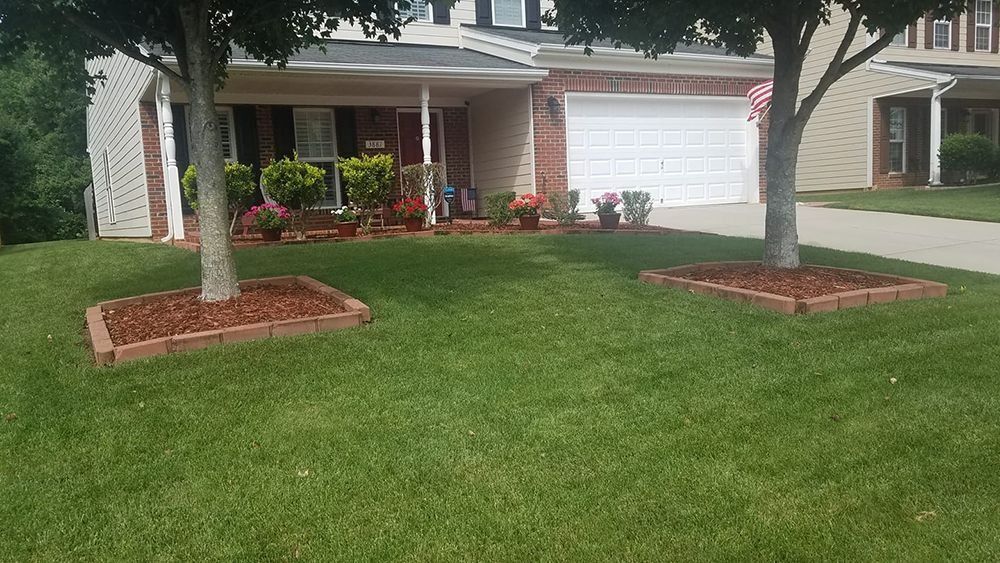 Lawn with two trees surrounded by brick edging. House with garage and porch in the background.