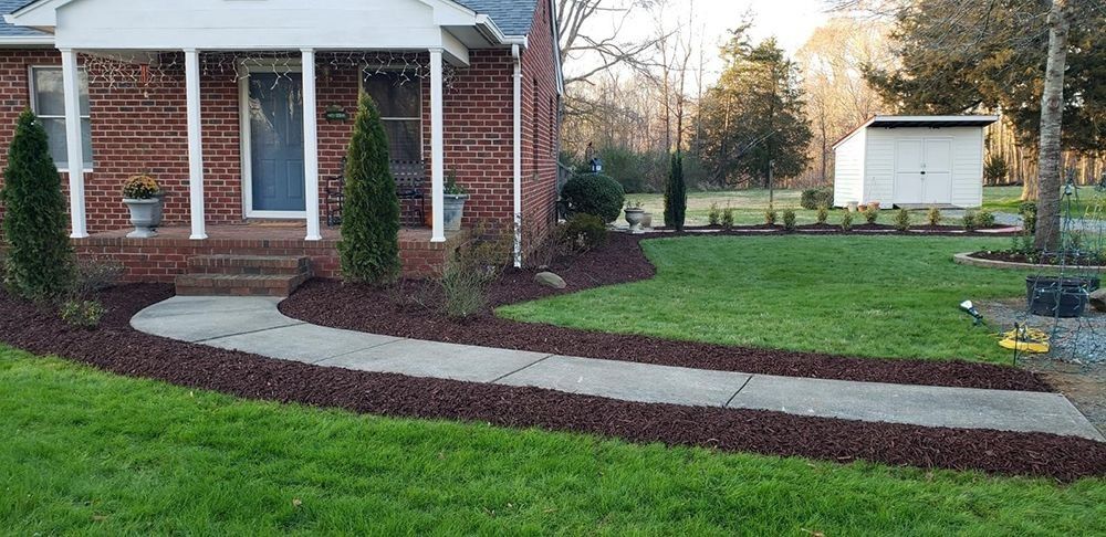 Brick house with white porch, curved walkway bordered by brown mulch, green lawn, and shed.