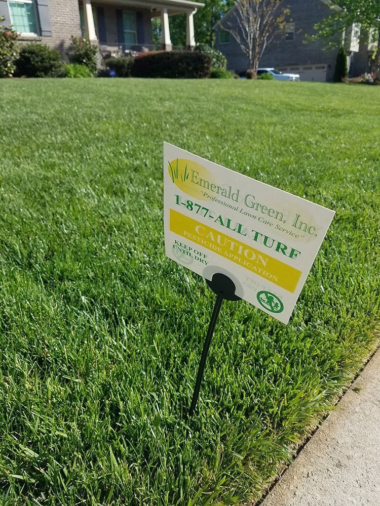 Green lawn with a caution sign from Emerald Green, Inc. near a sidewalk and house.