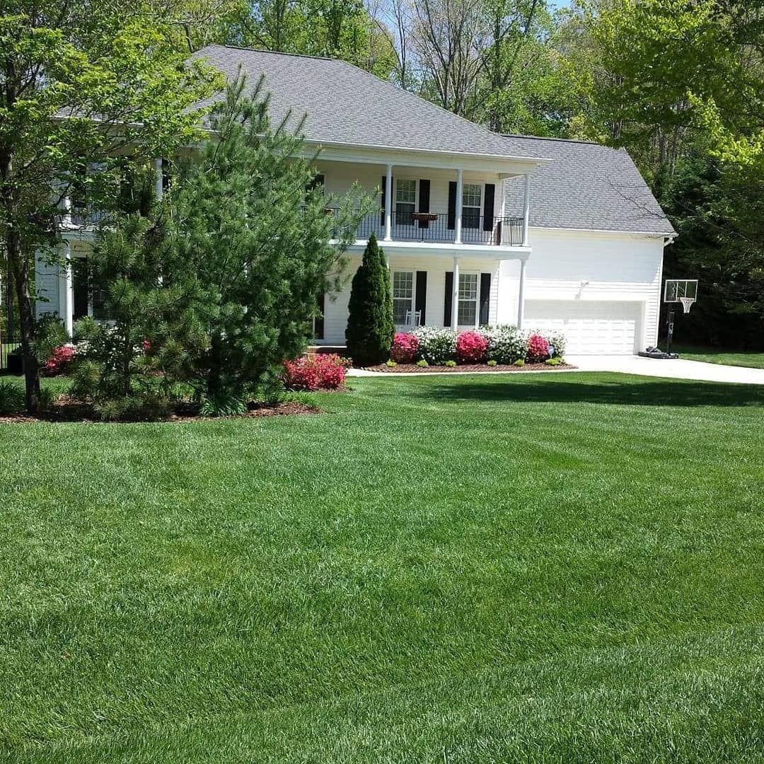 Two-story white house with green lawn, trees, and pink/red flowers; sunny day.