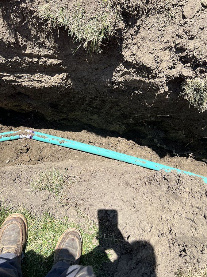 Trench with a blue PVC pipe running through it; person's boots and shadow visible.