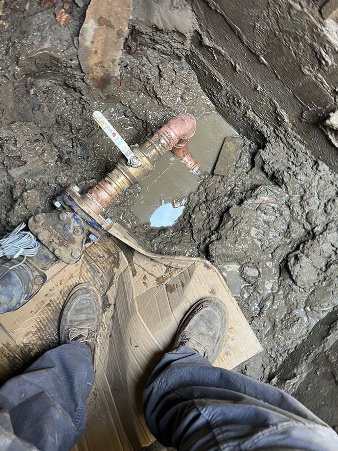 A person looking down at a water valve in a muddy pit. Brown shoes, cardboard, and bronze pipes are visible.