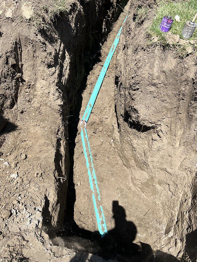 Trench dug in soil with a green pipe visible; shadow of a person's legs.