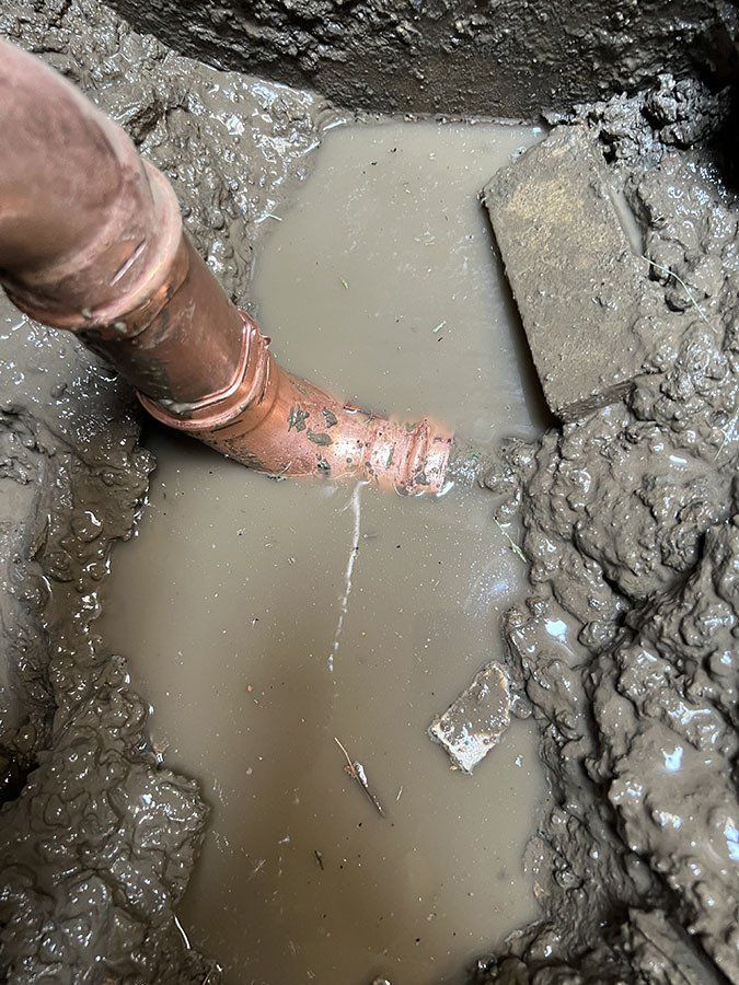 Copper pipe leaking water into muddy, dirt-filled hole, possibly a plumbing issue.
