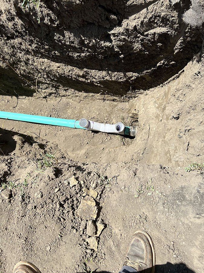 Person in work boots in a dirt trench, alongside a turquoise pipe and plumbing fitting.