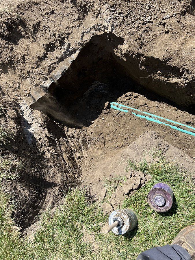 A trench dug in dirt reveals blue pipes; two metal fixtures lie on grass nearby.