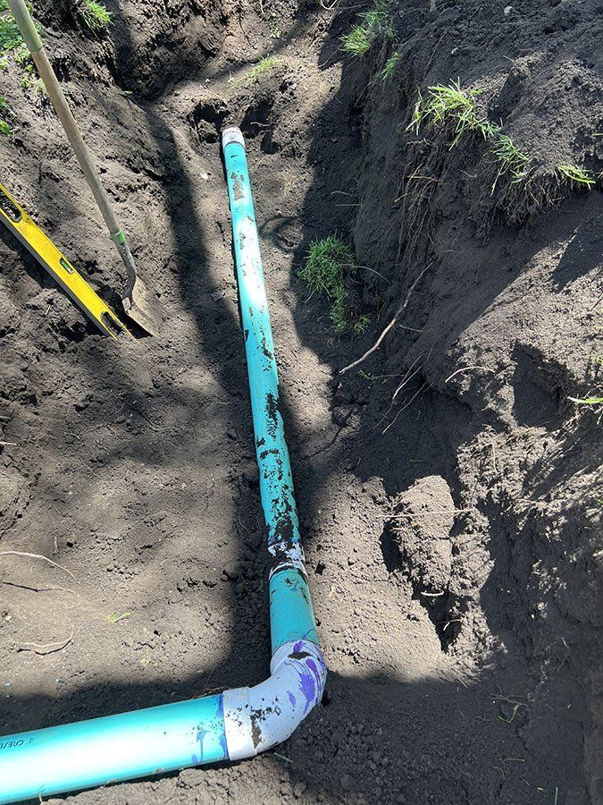 Turquoise PVC pipe in a trench, connected with white elbow fittings, partially buried in dirt.