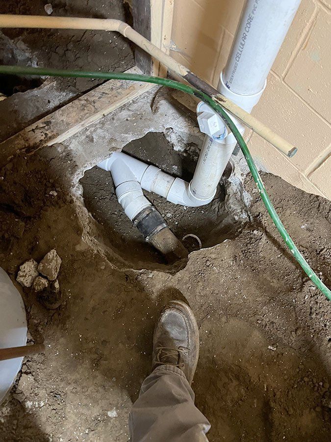 Plumbing pipes in a hole dug in dirt floor near a brick wall, with a person's foot visible.