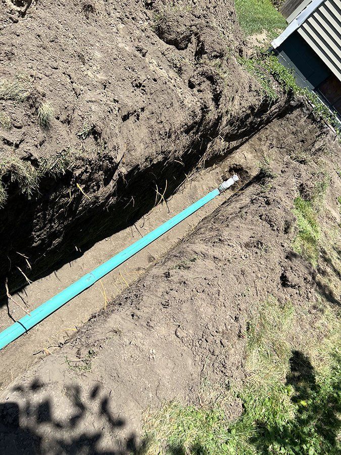 A trench dug in dirt holds a turquoise pipe, likely for plumbing, near a building's edge.