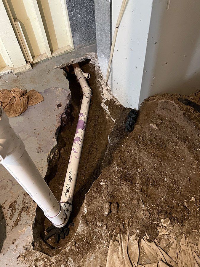 A partially buried PVC pipe in a trench next to a wall. Earth and concrete are visible.