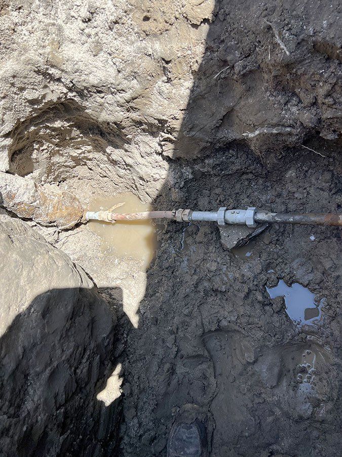 A broken water pipe in a dirt trench, with water present. Copper and plastic pipe.