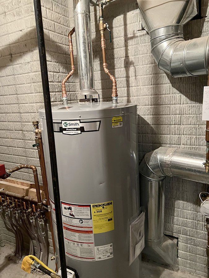 A water heater in a basement. It's a tall, gray cylinder with copper pipes and a metal flue against a brick wall.