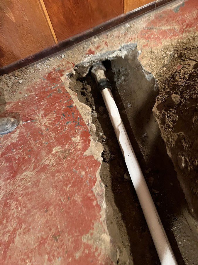 A trench in a concrete floor reveals a white pipe. The floor is red, and the walls are wood-paneled.