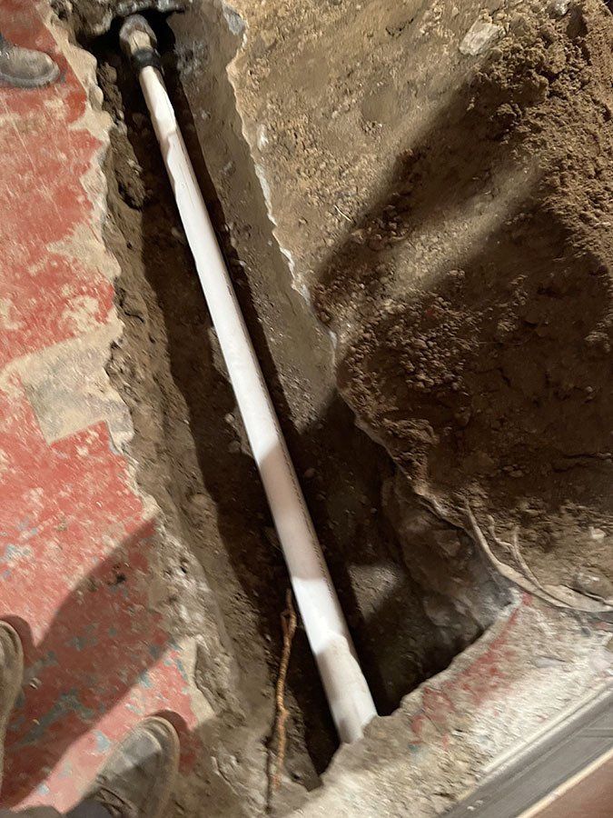 White pipe in a narrow trench dug in concrete and dirt; likely plumbing repair.