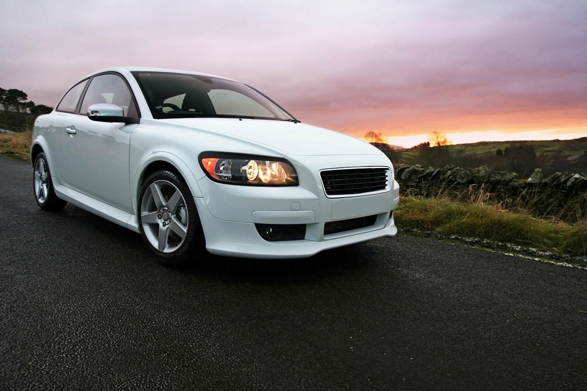 White Volvo C30 coupe parked on a wet road at sunset.
