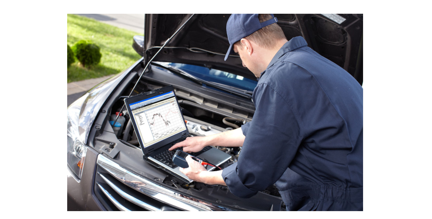 Mechanic using laptop to diagnose a car engine; open hood, outdoors, blue work clothes.