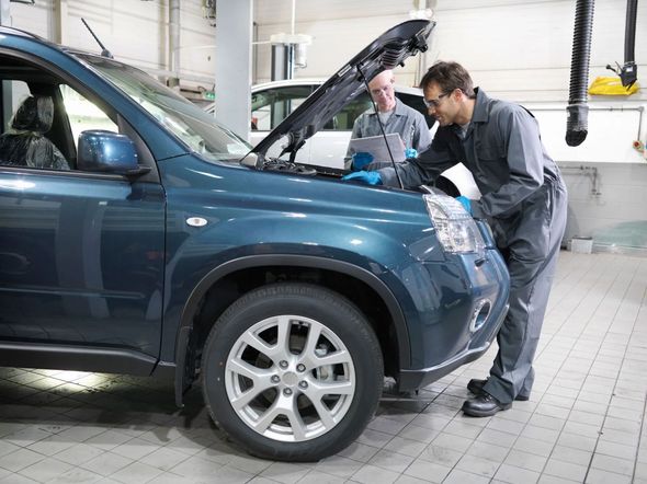 Two mechanics examining a blue car's engine in a garage. One uses a laptop.
