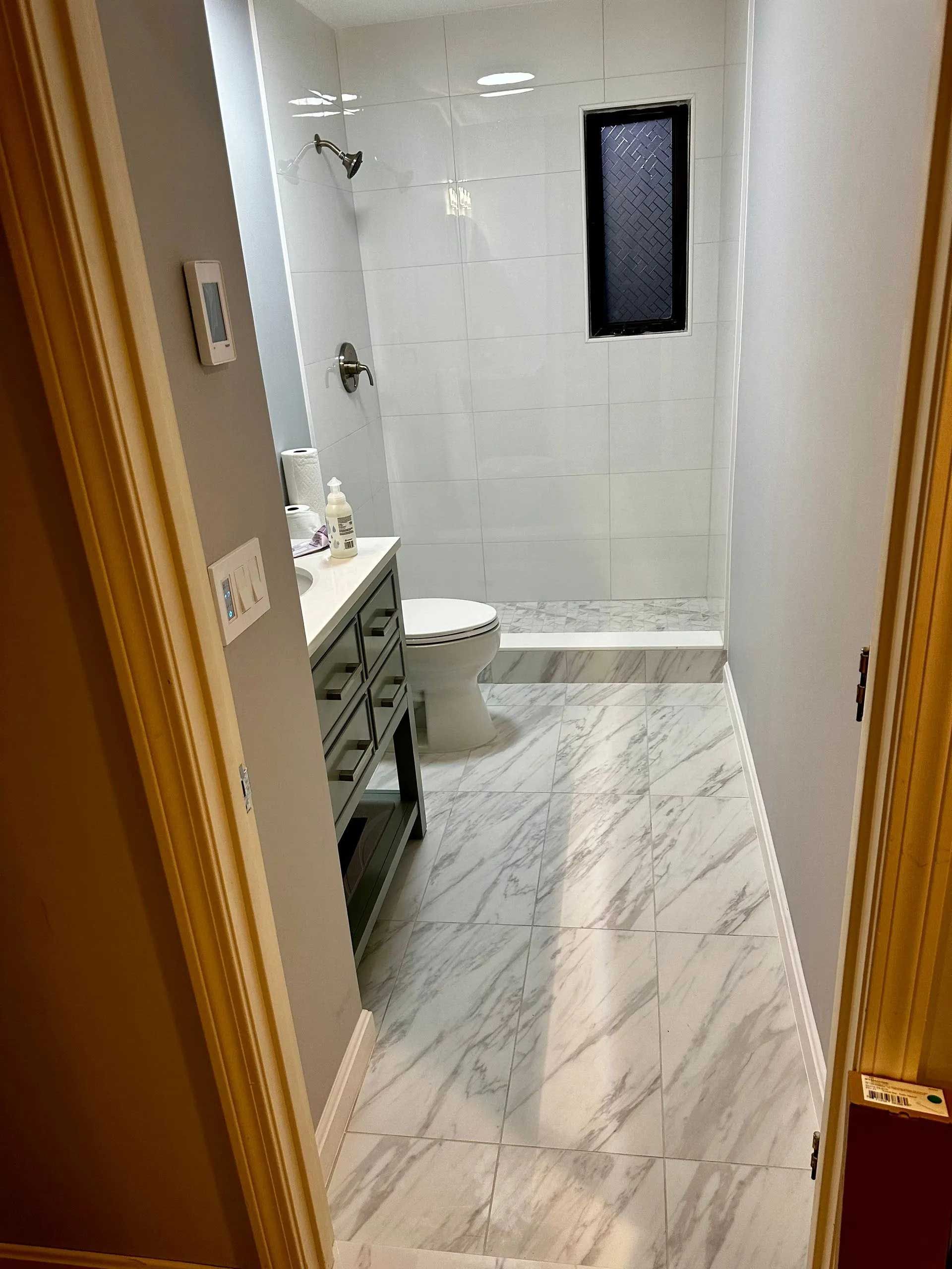 Narrow bathroom with marble-patterned floor and white tiled shower, gray vanity, and small window.