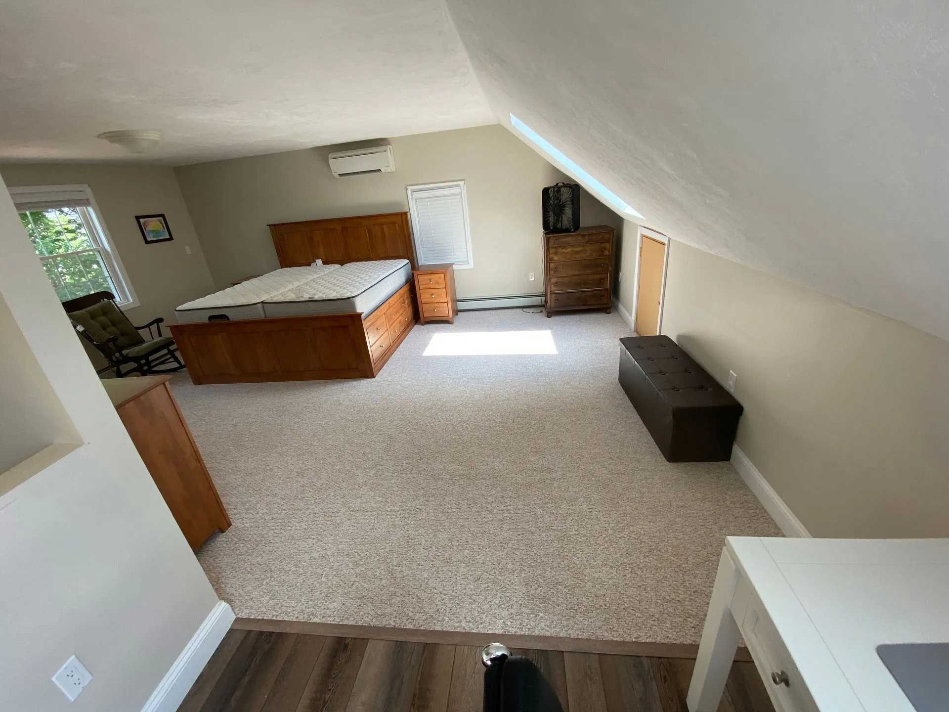 Bedroom with bed, dressers, a storage bench, desk, and chair, in a room with angled ceiling and neutral colors.