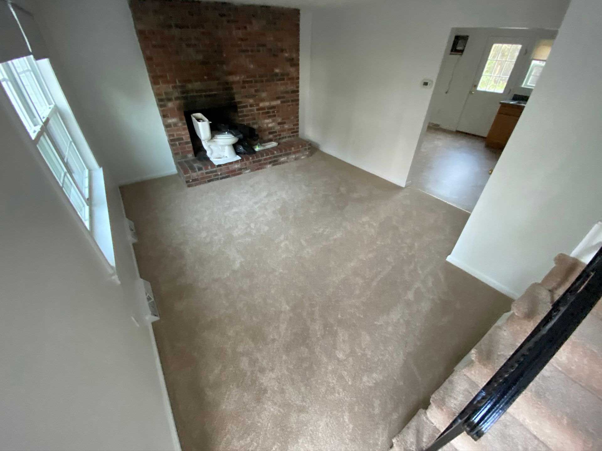 Living room with brick fireplace, beige carpet, stairs with a black railing, and a doorway to a kitchen.