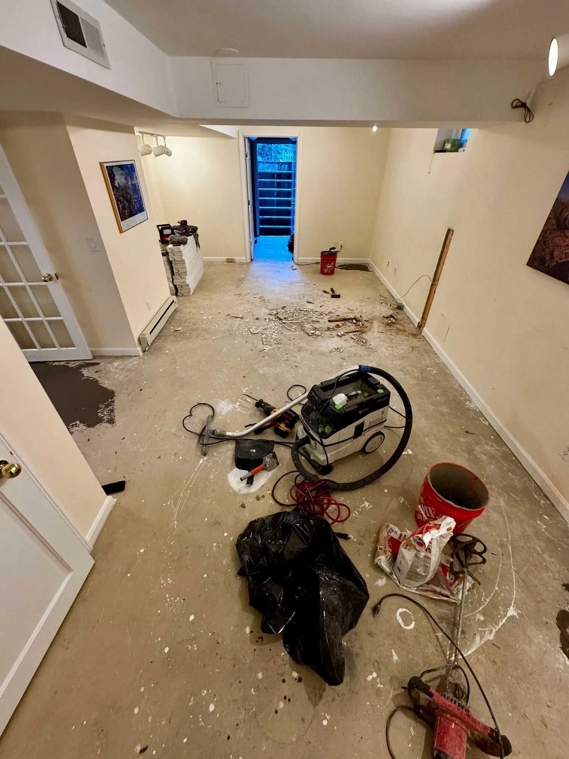 Basement room in disrepair with scattered debris and tools on the floor, doorway visible at the end.