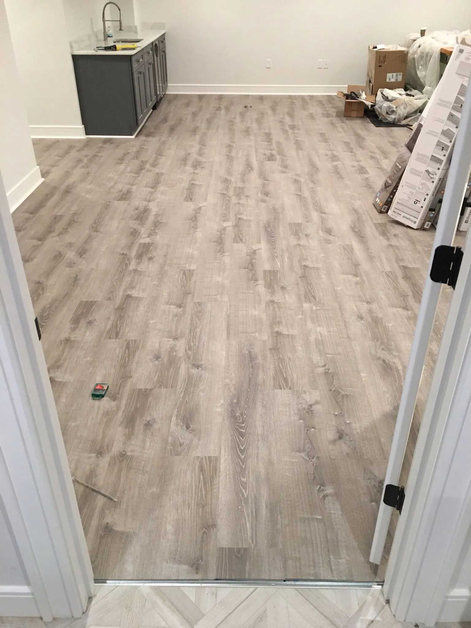 Room with light wood-look flooring. Gray cabinet and sink on the left. Doorway and construction materials.
