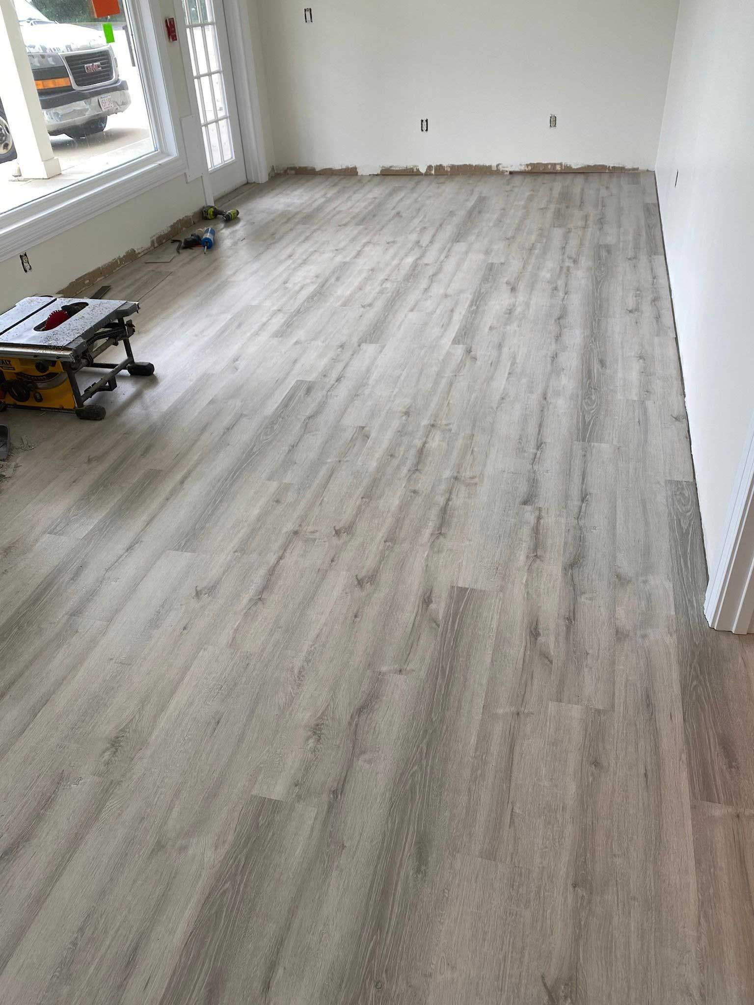 Newly installed gray wood-look flooring in a room with white walls. A saw is visible on the left.