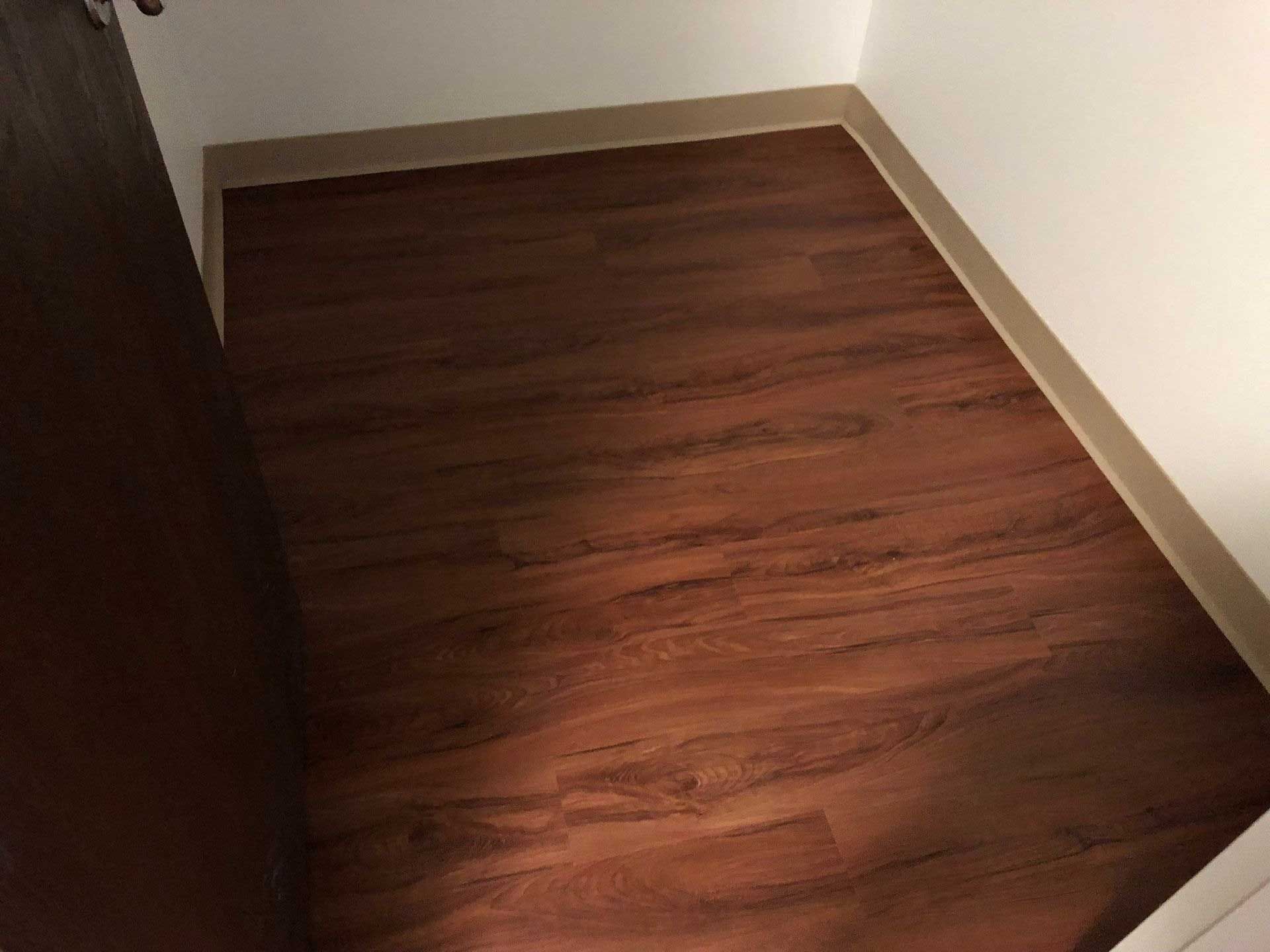 Empty small room with dark wood-look flooring and beige trim, brown door on left, white wall on right.