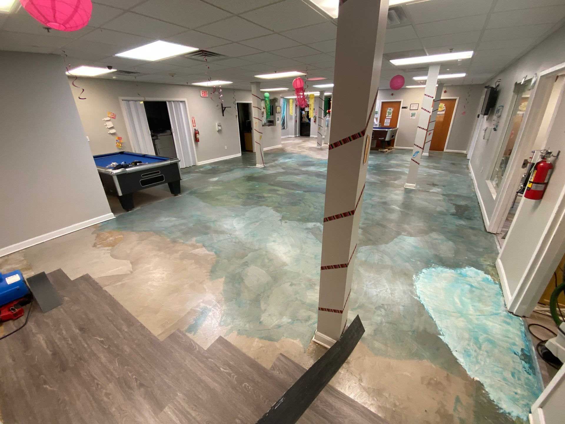 An empty event space with a pool table. Grey, blue and green paint on the floor.  Pink balloons hang.