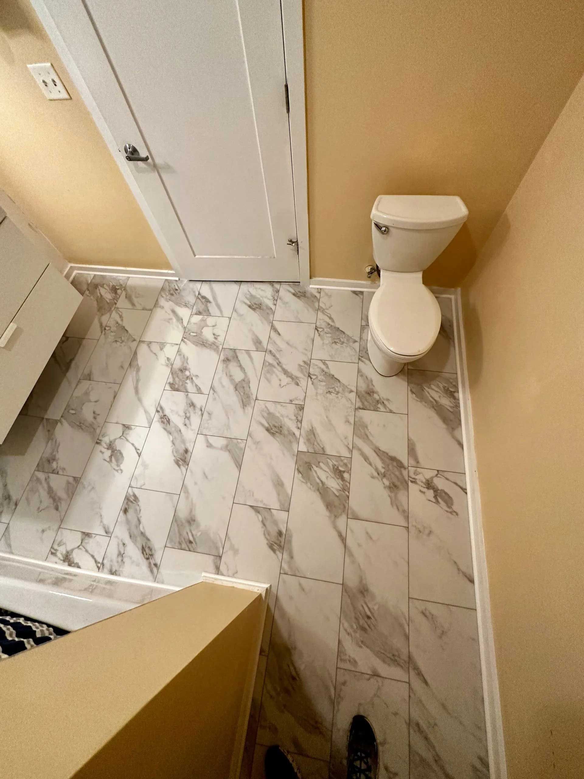 Small bathroom with marble-look flooring, white toilet, and closed white door. Yellow walls, angled perspective.