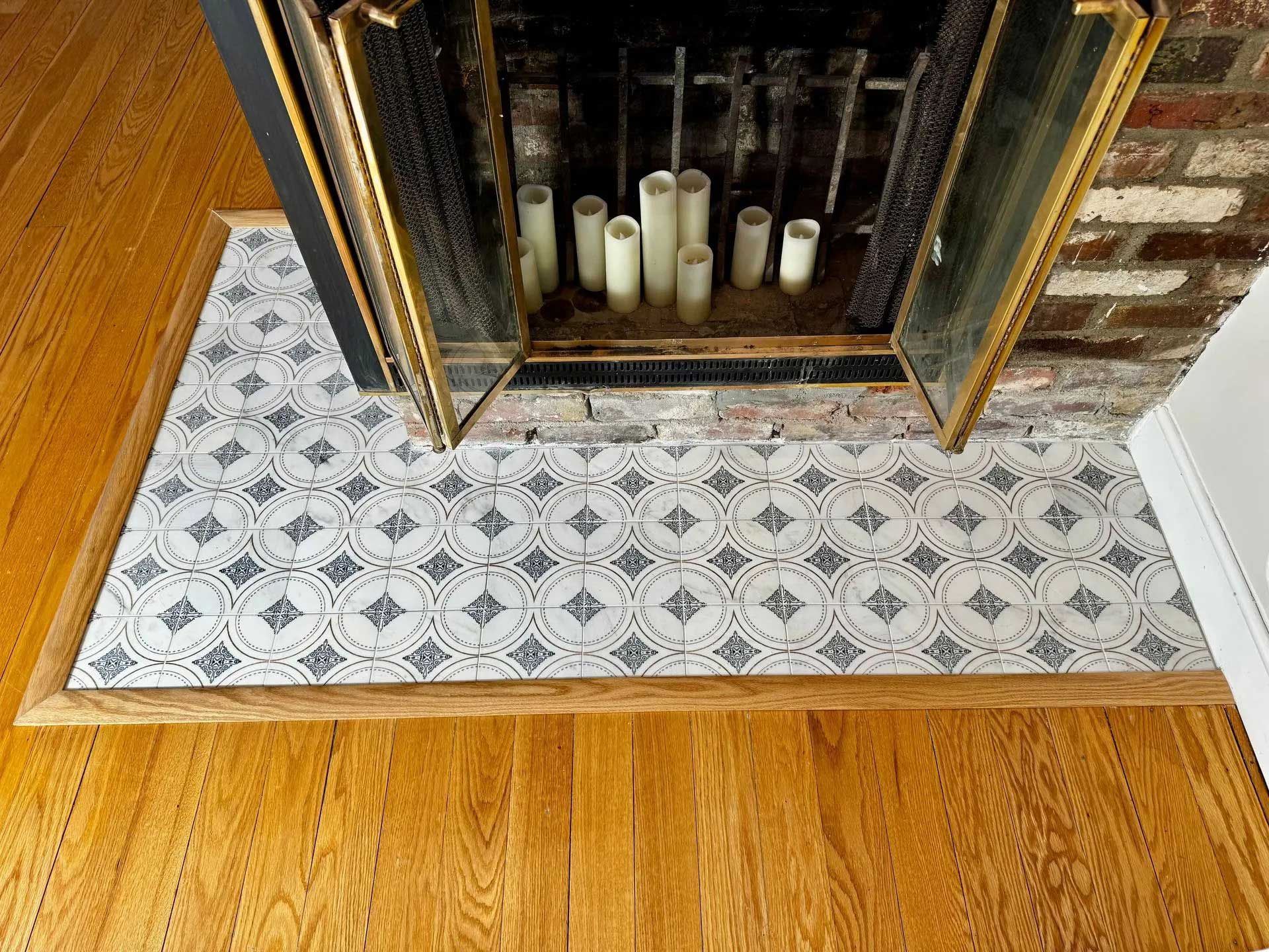 Fireplace with patterned tile hearth and gold-framed glass doors on wood flooring.