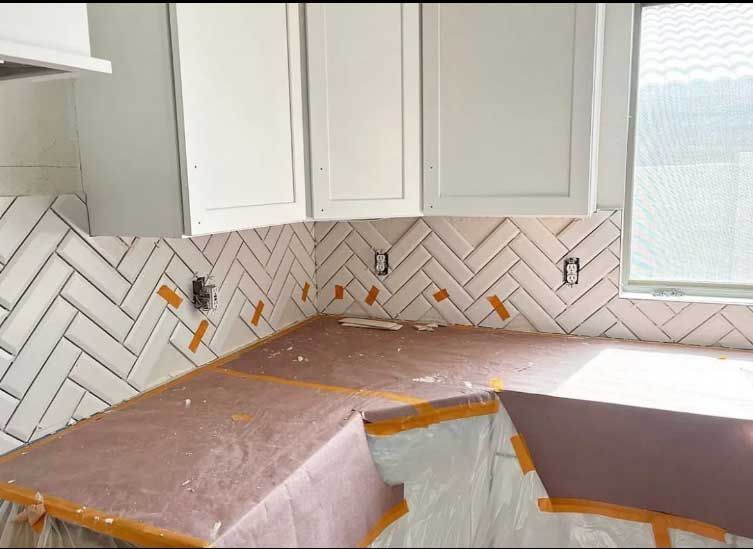 Kitchen backsplash with white herringbone tiles and upper white cabinets. Countertops are protected.