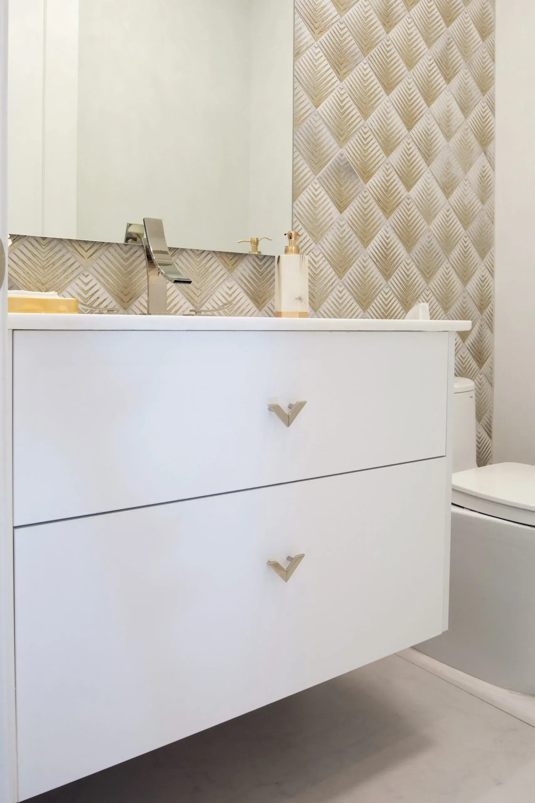 White bathroom vanity with gold handles beside a toilet and patterned wallpaper, seen in a mirror.