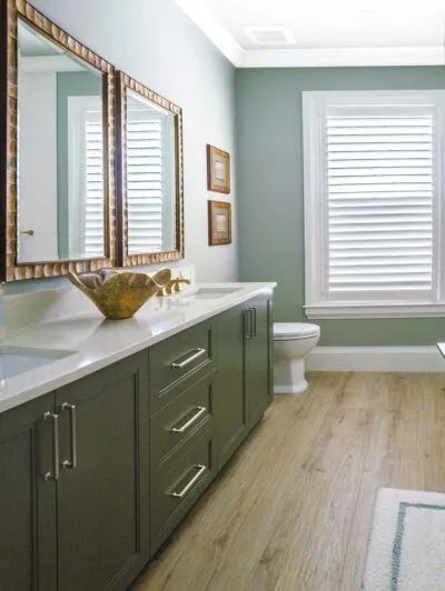 Modern bathroom with green vanity, large mirrors, white window blinds, and light wood flooring.