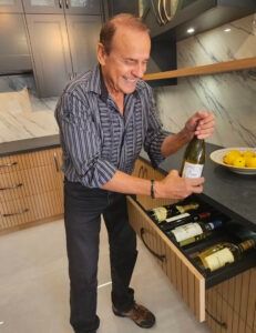 Man in a striped shirt opening a wine bottle beside an open kitchen drawer with wine bottles and a bowl of lemons