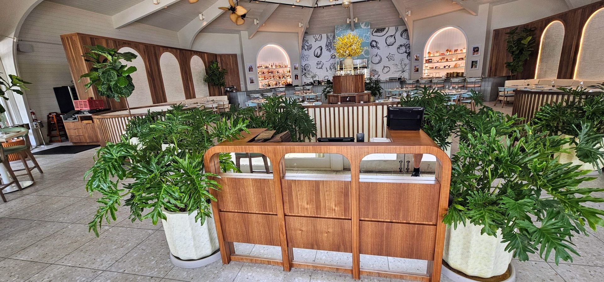 Bright indoor garden lounge with wooden seating, lush green plants, and arched windows in a spacious hall
