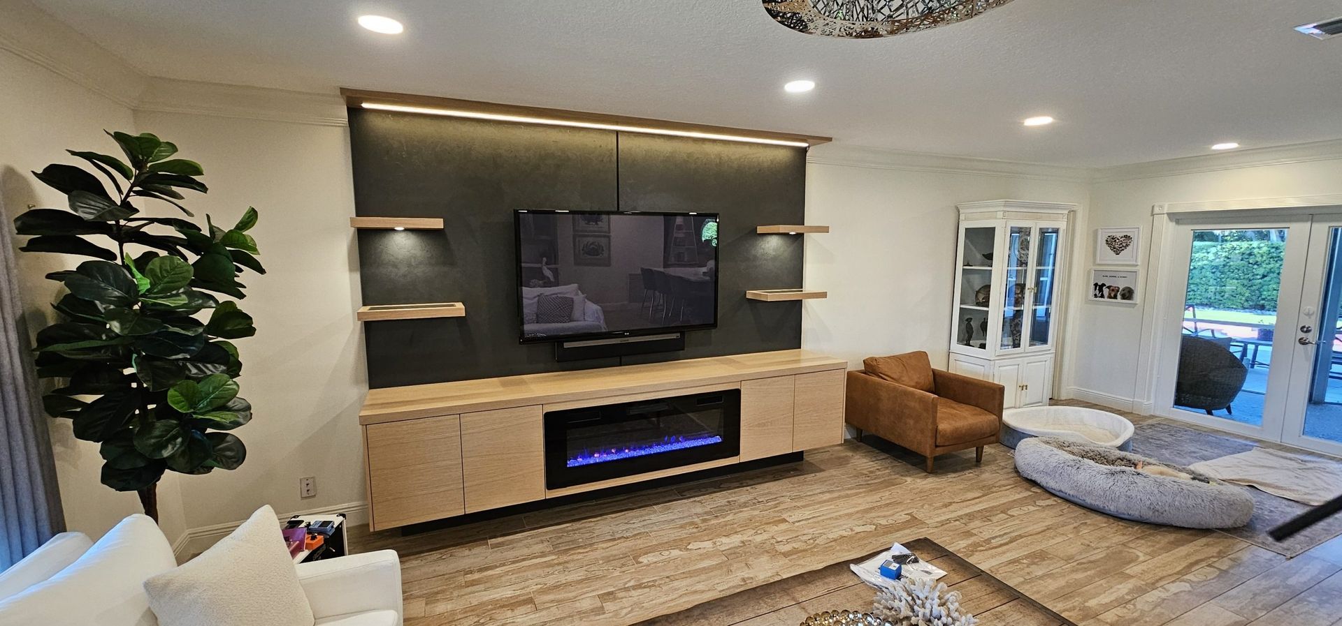 Modern living room with mounted TV, fireplace, tan sofa, large plant, and open glass doors to a patio.