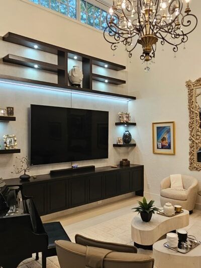Elegant living room with large TV, black shelving, cream chairs, glass coffee table, and chandelier on a high ceiling.