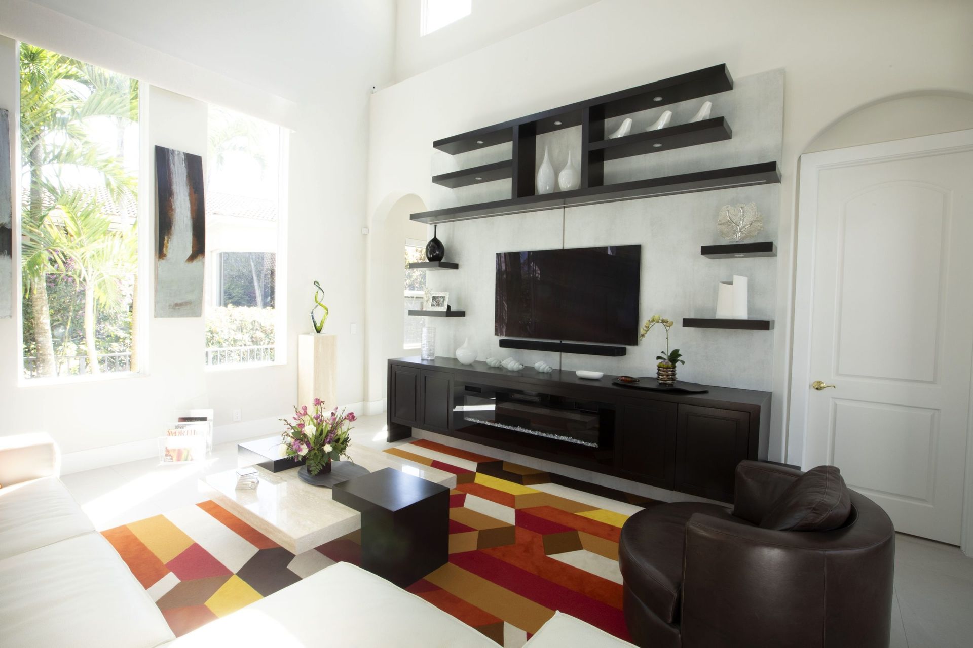 Bright modern living room with wall-mounted TV, black shelving, colorful rug, and large windows.