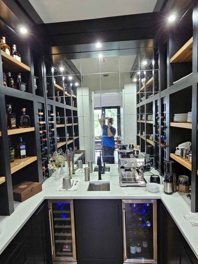 Narrow wine cellar with black shelves, bottles, mirrored back wall, and a person in blue reflected at the end