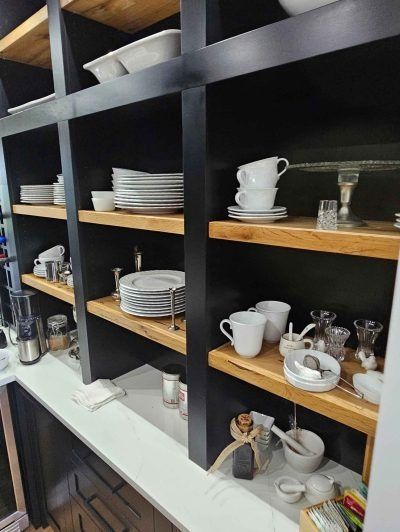 Black shelving with wooden shelves displaying stacked white dishes, cups, and glassware in a café or kitchen.