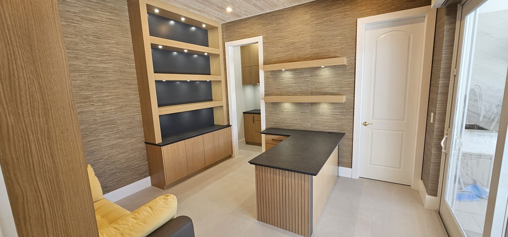 Modern kitchen with wood cabinetry, black countertop island, white door, and glass shelving near a window