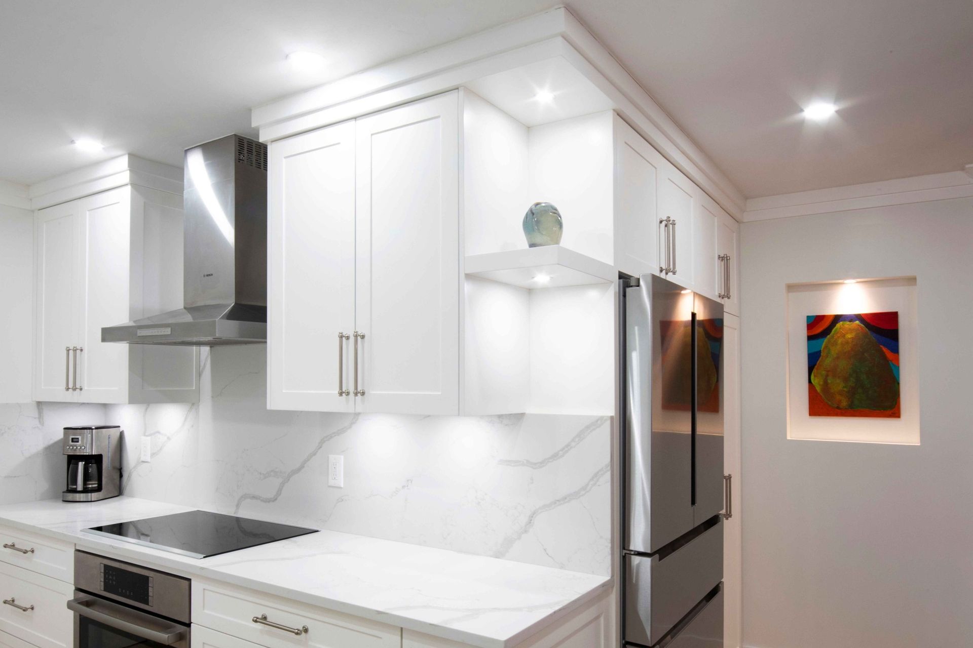 Modern white kitchen with marble counters, stainless appliances, and colorful wall art in the hallway