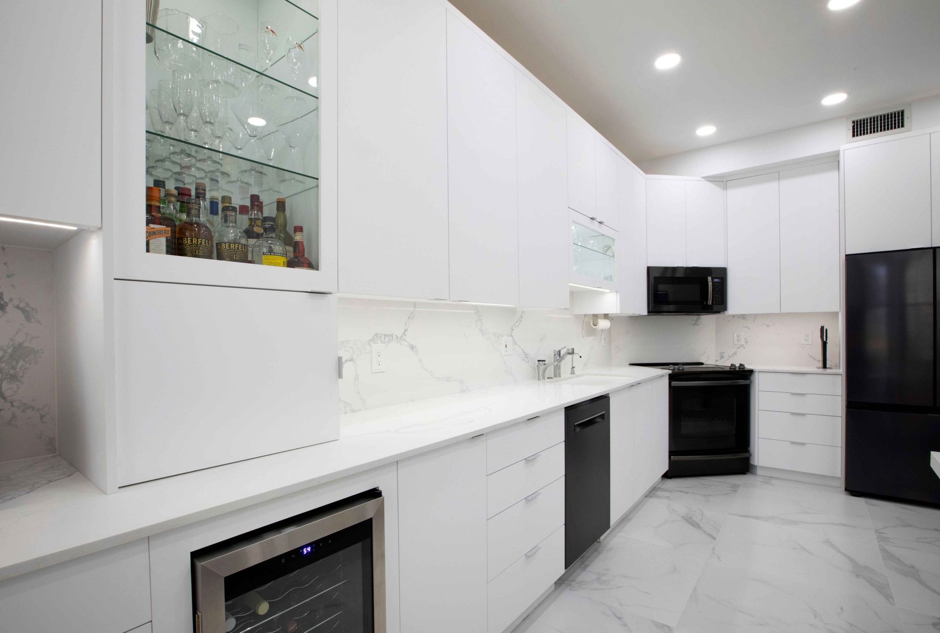 Modern white kitchen with marble floors, glossy cabinets, black appliances, and a glass display cabinet
