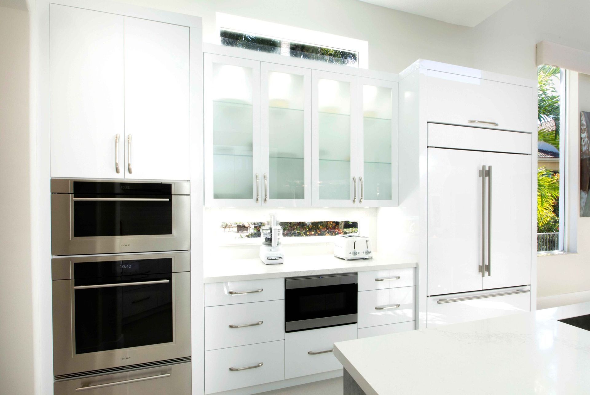 Bright white modern kitchen with stainless steel appliances, glass cabinets, and a large window