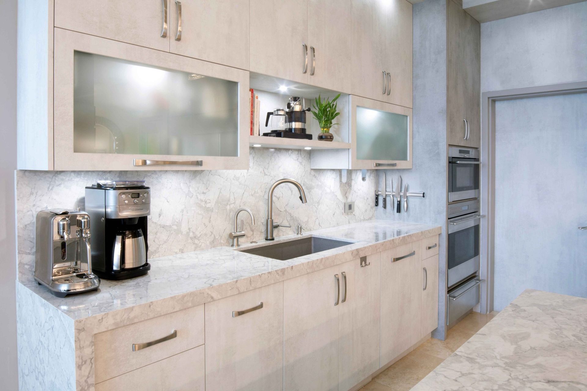 Modern light-wood kitchen with marble counters, stainless sink, coffee maker, and built-in oven