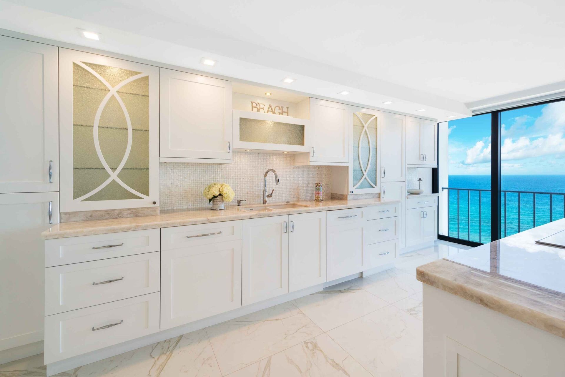 Bright white coastal kitchen with marble counters and ocean view through open balcony doors