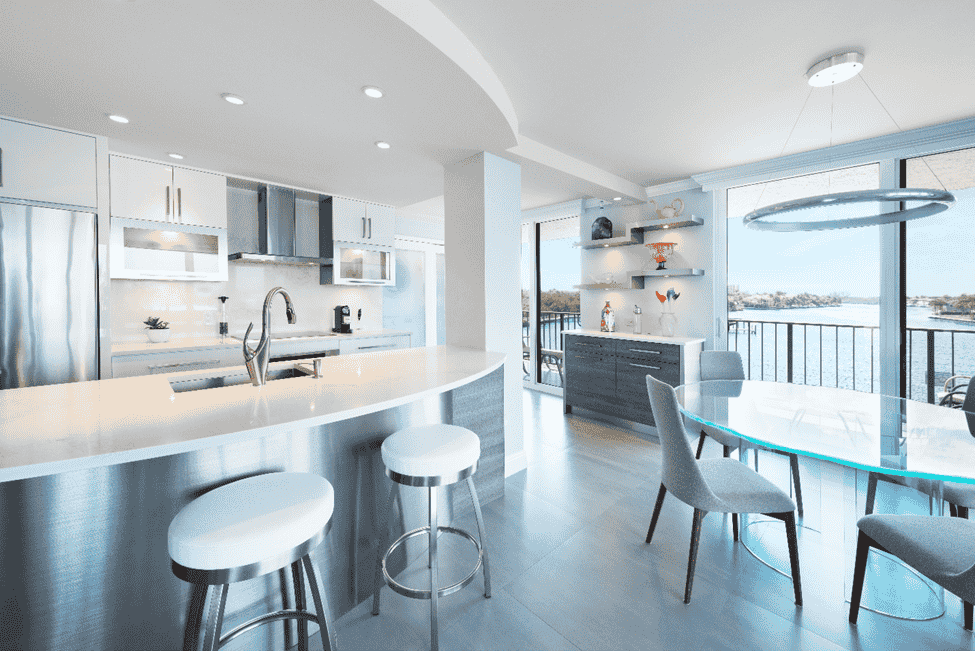 Bright modern kitchen and dining area with white cabinets, island stools, and large windows overlooking water