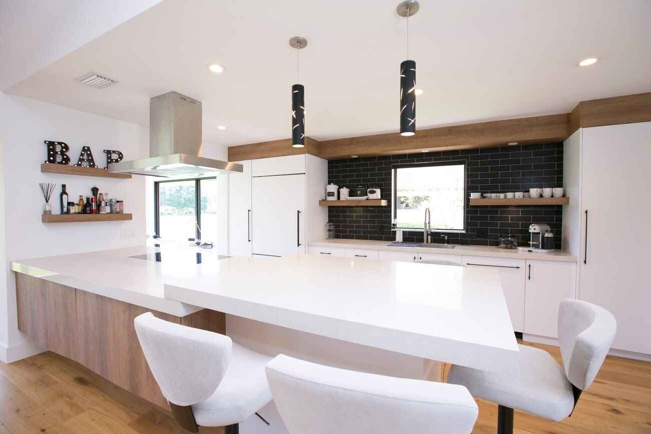Modern white kitchen with island, bar stools, wood accents, and black backsplash under pendant lights